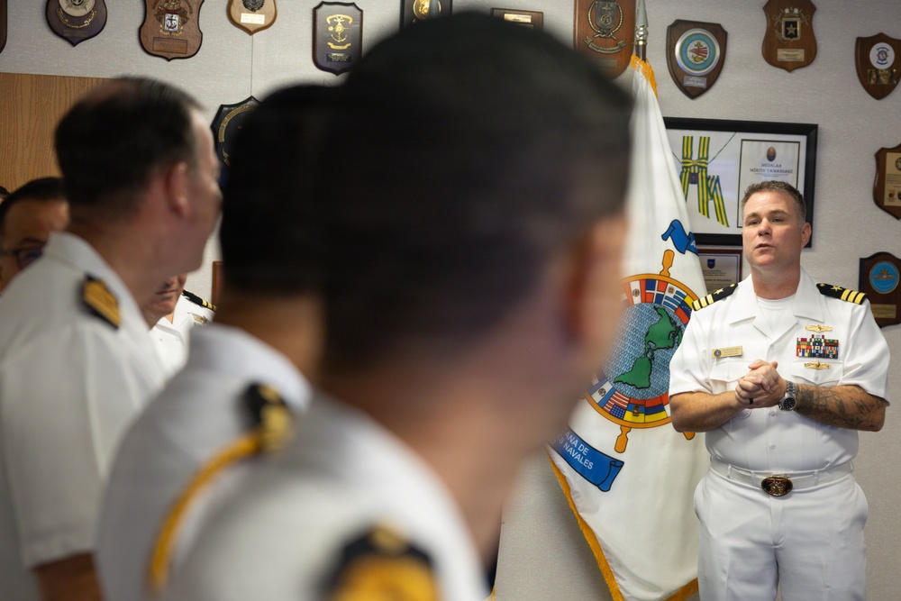 A naval officer in white dress uniform addresses a group of fellow officers in what appears to be an official meeting room or headquarters. The speaker stands in front of a ceremonial flag featuring a globe and ship's wheel design, surrounded by plaques and insignia on the walls. The setting suggests a multinational or inter-naval cooperative meeting.