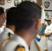 A naval officer in white dress uniform addresses a group of fellow officers in what appears to be an official meeting room or headquarters. The speaker stands in front of a ceremonial flag featuring a globe and ship's wheel design, surrounded by plaques and insignia on the walls. The setting suggests a multinational or inter-naval cooperative meeting.