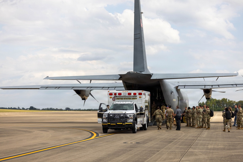 A United States Army ambulance is positioned in front of a large military transport aircraft, likely a C-130 Hercules, on a tarmac. A group of military personnel in camouflage uniforms are gathered near the rear cargo ramp of the aircraft. The scene appears to depict a military medical or logistics operation at an airfield.