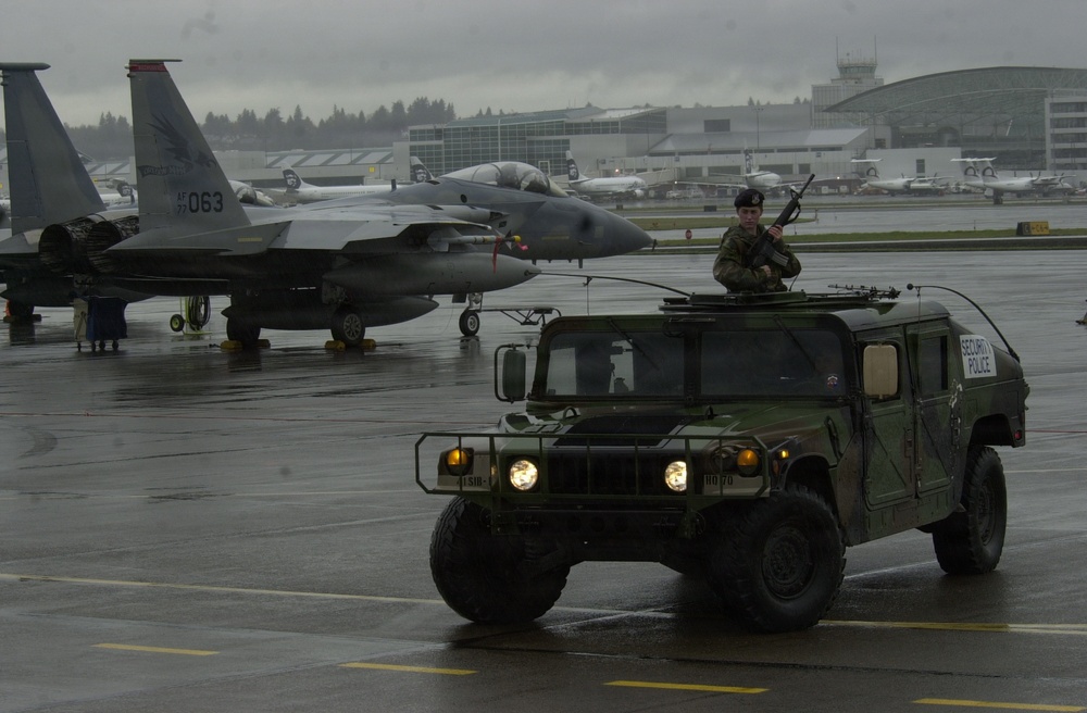 A military Security Police HMMWV (Humvee) patrols a wet airport tarmac during overcast, rainy conditions. An armed soldier stands through the roof hatch holding a rifle while the vehicle's headlights are illuminated. In the background, at least two F-15 Eagle fighter jets are parked on the flight line near what appears to be a joint civilian-military airfield.