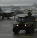 A military Security Police HMMWV (Humvee) patrols a wet airport tarmac during overcast, rainy conditions. An armed soldier stands through the roof hatch holding a rifle while the vehicle's headlights are illuminated. In the background, at least two F-15 Eagle fighter jets are parked on the flight line near what appears to be a joint civilian-military airfield.
