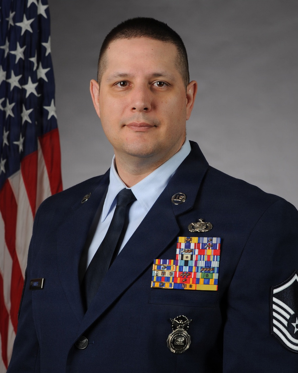 A formal military portrait of a United States Air Force enlisted member in dress blue uniform. The subject displays numerous service ribbons, badges, and rank insignia consistent with a senior non-commissioned officer. An American flag is visible in the background, typical of official military portrait settings.
