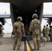Military personnel in camouflage uniforms are boarding or preparing to board a military transport aircraft via its rear cargo ramp. Two soldiers in the foreground wear flight helmets and tactical gear with backpacks, while a larger group of uniformed personnel stands to the right. A photographer or videographer documents the event from the left side of the frame.