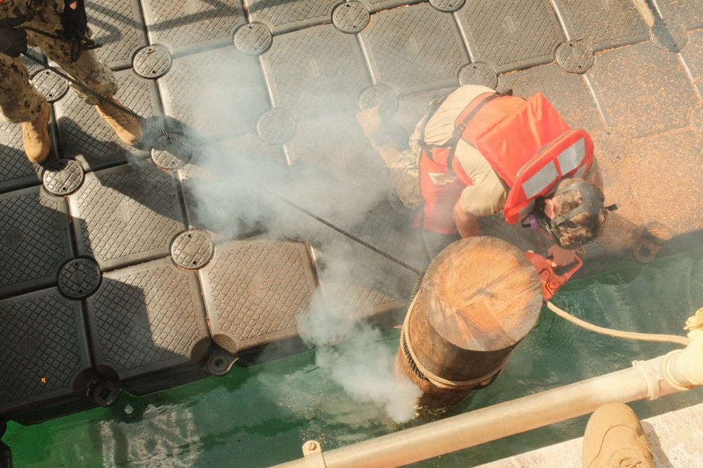 An aerial/overhead view of what appears to be a military or naval operation on a dock or vessel deck. A person wearing an orange life vest and helmet is working near the water's edge, where a large cylindrical object partially submerged in green water is emitting white smoke or steam. Additional personnel in military-style boots are visible at the edge of the frame.