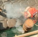 An aerial/overhead view of what appears to be a military or naval operation on a dock or vessel deck. A person wearing an orange life vest and helmet is working near the water's edge, where a large cylindrical object partially submerged in green water is emitting white smoke or steam. Additional personnel in military-style boots are visible at the edge of the frame.
