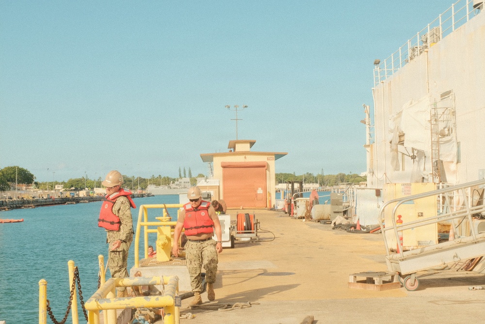 Two naval personnel wearing red life vests and camouflage uniforms are working on a military pier or dock facility. A large naval vessel is moored alongside the pier on the right side of the image. The setting appears to be a tropical naval base, likely Pearl Harbor, Hawaii, based on the vegetation and architectural style.