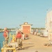 Two naval personnel wearing red life vests and camouflage uniforms are working on a military pier or dock facility. A large naval vessel is moored alongside the pier on the right side of the image. The setting appears to be a tropical naval base, likely Pearl Harbor, Hawaii, based on the vegetation and architectural style.
