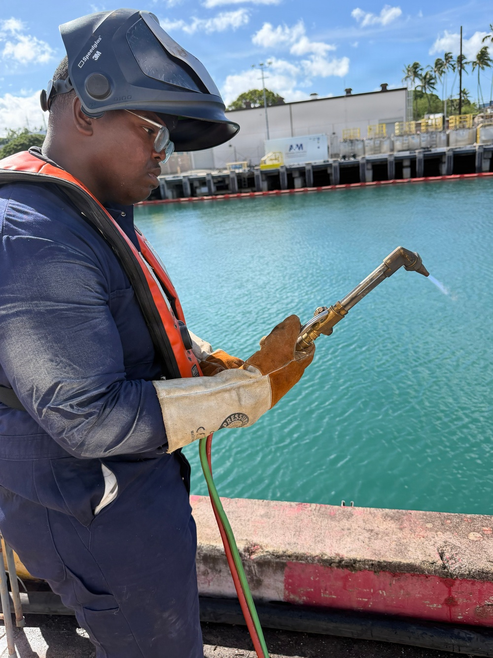 A worker is positioned near a waterway, holding an oxy-fuel cutting or welding torch with dual hoses (red and green, indicating oxygen and fuel gas). He is equipped with full personal protective equipment including a 3M Speedglas welding helmet, safety glasses, leather welding gloves, a blue work uniform, and an orange inflatable life vest. The setting appears to be a maritime industrial facility, likely a shipyard or port, with turquoise water and dock structures visible in the background.