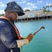 A worker is positioned near a waterway, holding an oxy-fuel cutting or welding torch with dual hoses (red and green, indicating oxygen and fuel gas). He is equipped with full personal protective equipment including a 3M Speedglas welding helmet, safety glasses, leather welding gloves, a blue work uniform, and an orange inflatable life vest. The setting appears to be a maritime industrial facility, likely a shipyard or port, with turquoise water and dock structures visible in the background.