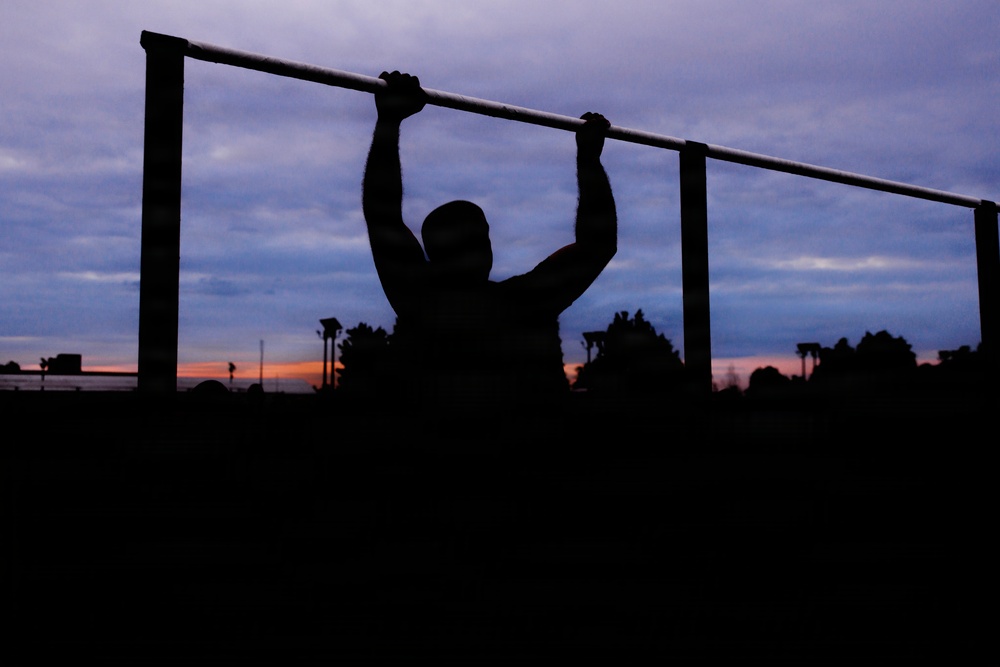 A silhouetted figure is performing pull-ups on an outdoor exercise bar during twilight or early dawn. The dramatic sky displays a gradient of deep blue, purple, and orange hues near the horizon. The scene appears to be set at an outdoor fitness area or park.