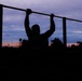 A silhouetted figure is performing pull-ups on an outdoor exercise bar during twilight or early dawn. The dramatic sky displays a gradient of deep blue, purple, and orange hues near the horizon. The scene appears to be set at an outdoor fitness area or park.