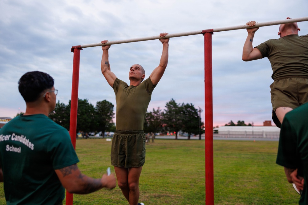 The image shows individuals performing pull-ups on an outdoor red metal pull-up bar structure during what appears to be early morning or dusk. At least one person on the left is wearing a green shirt with 'Officer Candidates School' text visible, indicating this is a U.S. Marine Corps Officer Candidates School (OCS) physical fitness assessment or training session. The setting is an open grassy field with trees and buildings visible in the background.