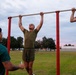 The image shows individuals performing pull-ups on an outdoor red metal pull-up bar structure during what appears to be early morning or dusk. At least one person on the left is wearing a green shirt with 'Officer Candidates School' text visible, indicating this is a U.S. Marine Corps Officer Candidates School (OCS) physical fitness assessment or training session. The setting is an open grassy field with trees and buildings visible in the background.