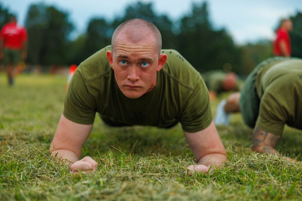 A close-up photograph of a young male in military-style olive green physical training attire performing a low plank or push-up position on a grass field. Multiple individuals in similar uniforms are visible in the background, suggesting a group physical training exercise. The setting appears to be an outdoor military or training facility.