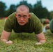 A close-up photograph of a young male in military-style olive green physical training attire performing a low plank or push-up position on a grass field. Multiple individuals in similar uniforms are visible in the background, suggesting a group physical training exercise. The setting appears to be an outdoor military or training facility.