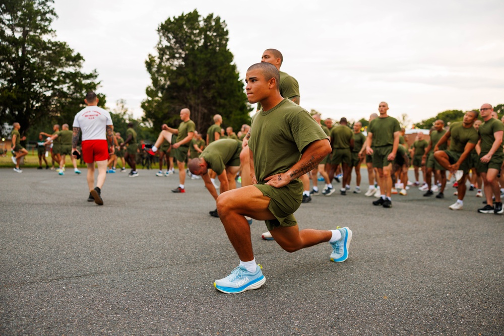 A large group of military personnel in olive green PT (physical training) uniforms are conducting an outdoor exercise session on a paved surface. The foreground shows a service member performing a lunge stretch, with dozens of others visible in various stretching positions behind them. An instructor in a white shirt and red shorts is visible supervising the formation in the background.