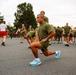 A large group of military personnel in olive green PT (physical training) uniforms are conducting an outdoor exercise session on a paved surface. The foreground shows a service member performing a lunge stretch, with dozens of others visible in various stretching positions behind them. An instructor in a white shirt and red shorts is visible supervising the formation in the background.