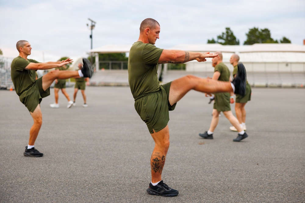 A group of military personnel in matching olive green PT (physical training) uniforms are performing synchronized high-knee stretching exercises on an outdoor paved surface. The foreground features a muscular male with a visible leg tattoo leading the exercise formation. The setting appears to be a military base or training facility with structures and trees visible in the background.