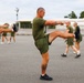 A group of military personnel in matching olive green PT (physical training) uniforms are performing synchronized high-knee stretching exercises on an outdoor paved surface. The foreground features a muscular male with a visible leg tattoo leading the exercise formation. The setting appears to be a military base or training facility with structures and trees visible in the background.