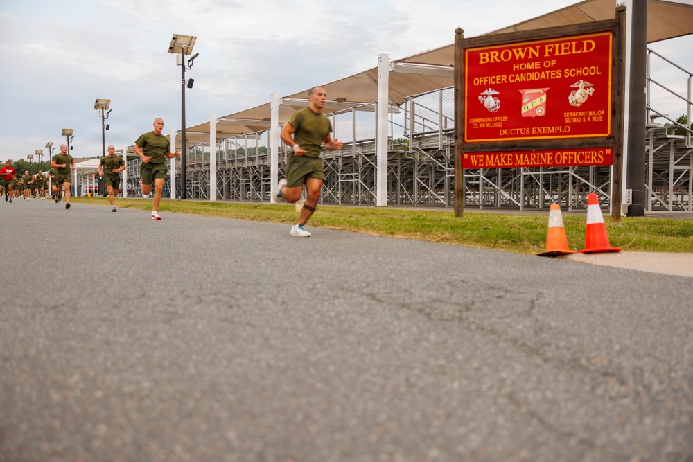 A group of military candidates in olive green PT (physical training) uniforms are running along a paved road at Brown Field, home of the Marine Corps Officer Candidates School (OCS). A prominent red sign in the foreground reads 'BROWN FIELD - HOME OF OFFICER CANDIDATES SCHOOL' with the motto 'DUCTUS EXEMPLO' and the phrase 'WE MAKE MARINE OFFICERS'. The scene captures a physical fitness run with multiple candidates stretching back along the route.