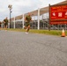 A group of military candidates in olive green PT (physical training) uniforms are running along a paved road at Brown Field, home of the Marine Corps Officer Candidates School (OCS). A prominent red sign in the foreground reads 'BROWN FIELD - HOME OF OFFICER CANDIDATES SCHOOL' with the motto 'DUCTUS EXEMPLO' and the phrase 'WE MAKE MARINE OFFICERS'. The scene captures a physical fitness run with multiple candidates stretching back along the route.