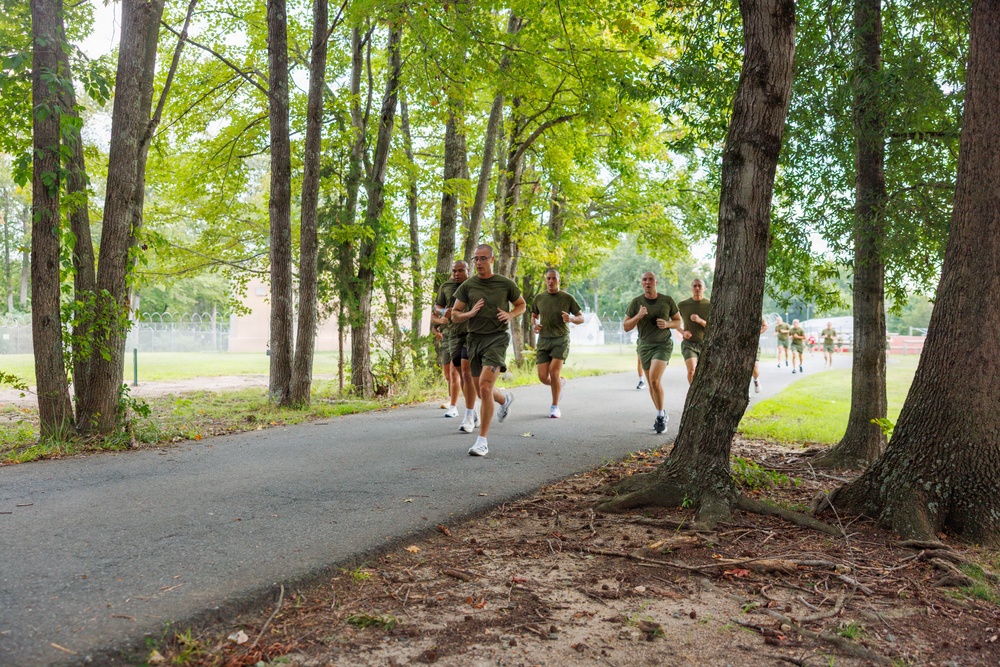 A group of military personnel in uniform physical training (PT) gear are running along a paved path lined with tall trees. They are wearing matching olive/dark green t-shirts and shorts with white athletic shoes, consistent with military PT uniforms. The setting appears to be a military base or installation, with fencing and open fields visible in the background.