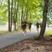 A group of military personnel in uniform physical training (PT) gear are running along a paved path lined with tall trees. They are wearing matching olive/dark green t-shirts and shorts with white athletic shoes, consistent with military PT uniforms. The setting appears to be a military base or installation, with fencing and open fields visible in the background.