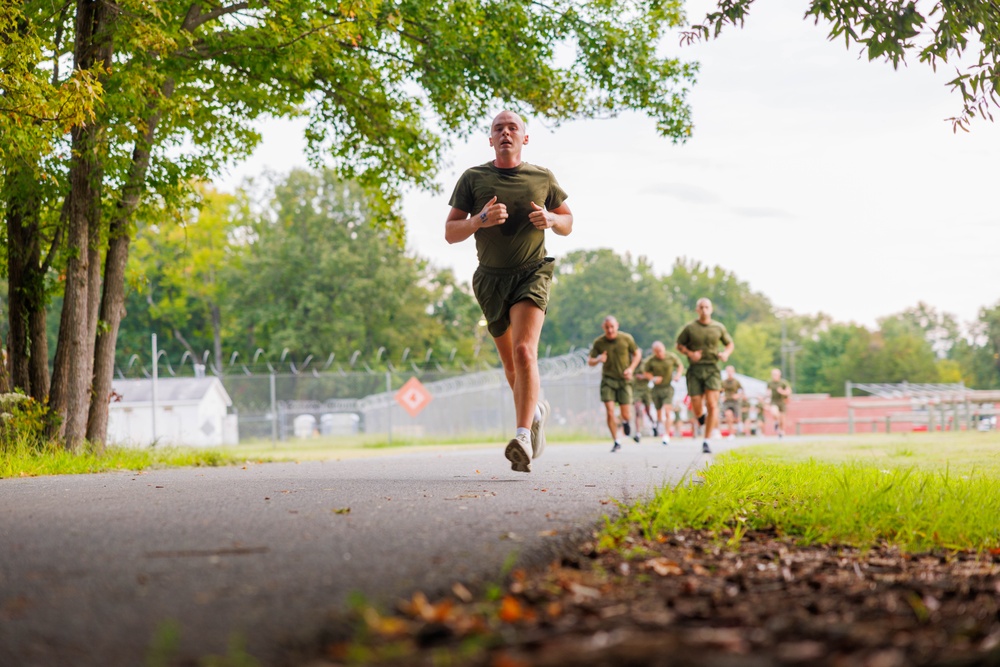 A group of individuals in matching olive green military-style physical training (PT) uniforms are running along a paved path. The lead runner is in sharp focus in the foreground, with several others visible in the background. The setting appears to be a military installation or training facility, evidenced by fencing, a track, and structured grounds.