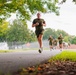 A group of individuals in matching olive green military-style physical training (PT) uniforms are running along a paved path. The lead runner is in sharp focus in the foreground, with several others visible in the background. The setting appears to be a military installation or training facility, evidenced by fencing, a track, and structured grounds.