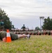A large group of military personnel in olive drab uniforms are performing push-ups in formation on a grass field. Instructors or supervisors in matching uniforms stand upright observing the exercise. The scene appears to be a physical training session at a military installation, with rows of recruits or trainees extending into the background.
