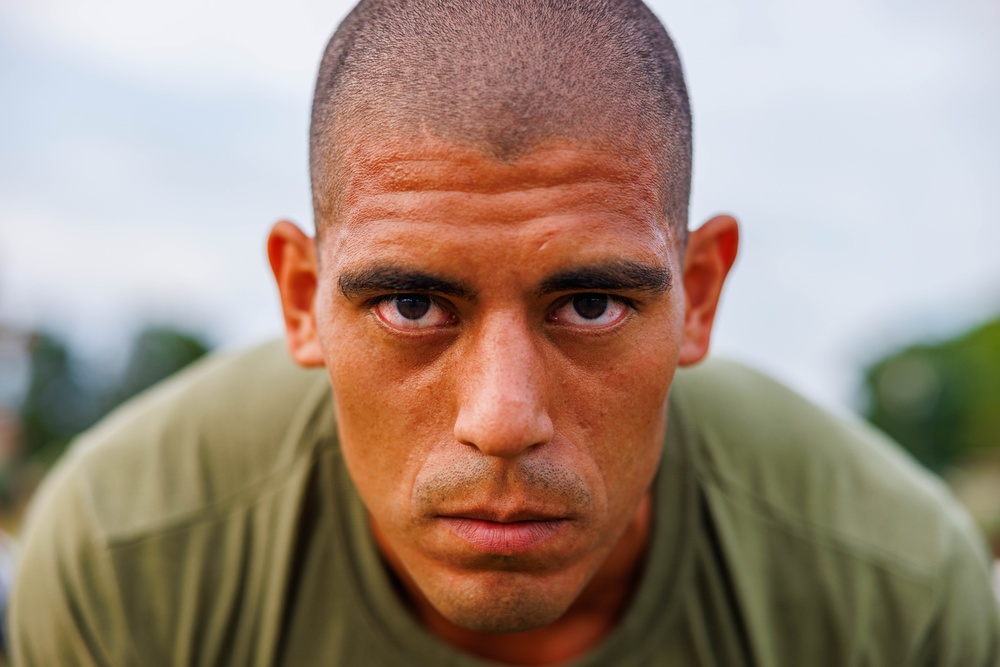 A close-up portrait of a young adult male with a shaved head, flushed and sweating face, staring directly into the camera with an intense expression. He is wearing an olive/army green t-shirt, suggesting possible military or athletic context. The background is blurred with hints of greenery and an overcast sky.