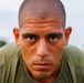 A close-up portrait of a young adult male with a shaved head, flushed and sweating face, staring directly into the camera with an intense expression. He is wearing an olive/army green t-shirt, suggesting possible military or athletic context. The background is blurred with hints of greenery and an overcast sky.