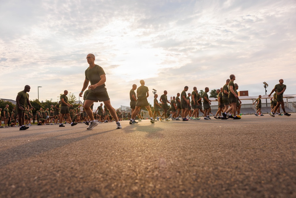 A large group of military personnel in olive green PT (physical training) uniforms are running or jogging in formation on a paved surface. The image is taken from a low ground-level angle during early morning or late afternoon, with the sun creating a strong backlight effect. The scene appears to be a organized military physical fitness run on a base or training facility.