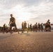 A large group of military personnel in olive green PT (physical training) uniforms are running or jogging in formation on a paved surface. The image is taken from a low ground-level angle during early morning or late afternoon, with the sun creating a strong backlight effect. The scene appears to be a organized military physical fitness run on a base or training facility.