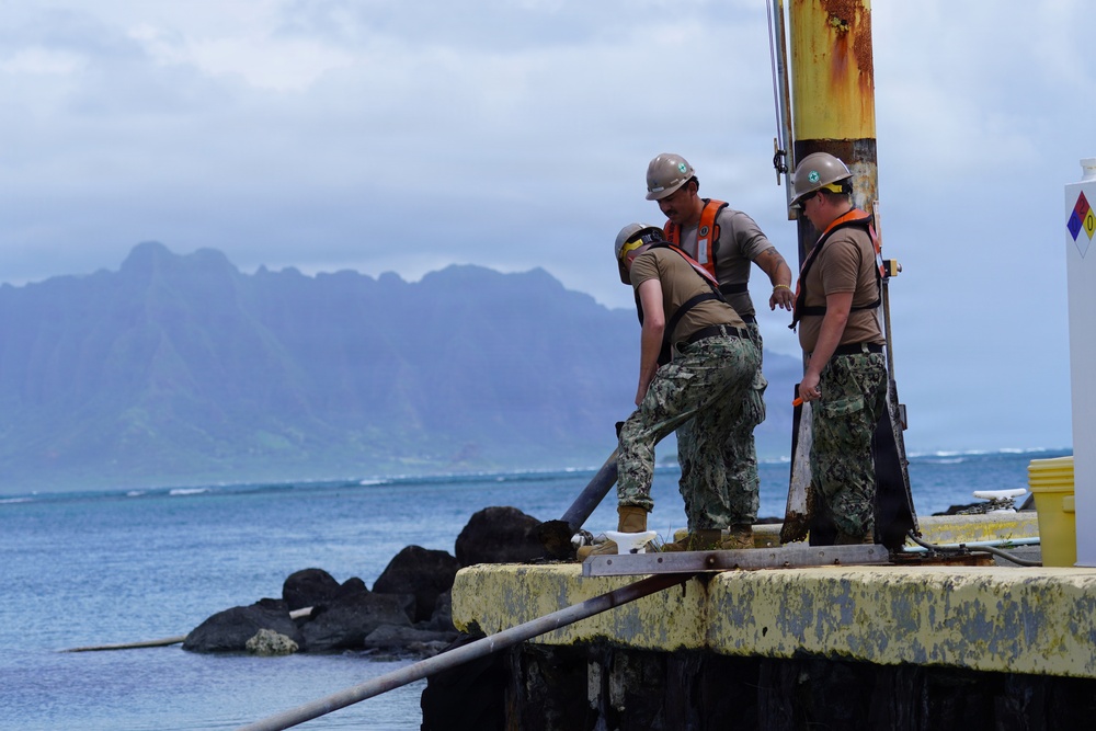 Three workers in military-style uniforms and hard hats are performing maintenance or construction work on a weathered pier or dock structure. They appear to be handling a pipe or hose near a rusted metal pole. The scene is set against a coastal backdrop with rocky shoreline, blue ocean water, and dramatic mountainous terrain under an overcast sky.