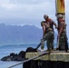 Three workers in military-style uniforms and hard hats are performing maintenance or construction work on a weathered pier or dock structure. They appear to be handling a pipe or hose near a rusted metal pole. The scene is set against a coastal backdrop with rocky shoreline, blue ocean water, and dramatic mountainous terrain under an overcast sky.