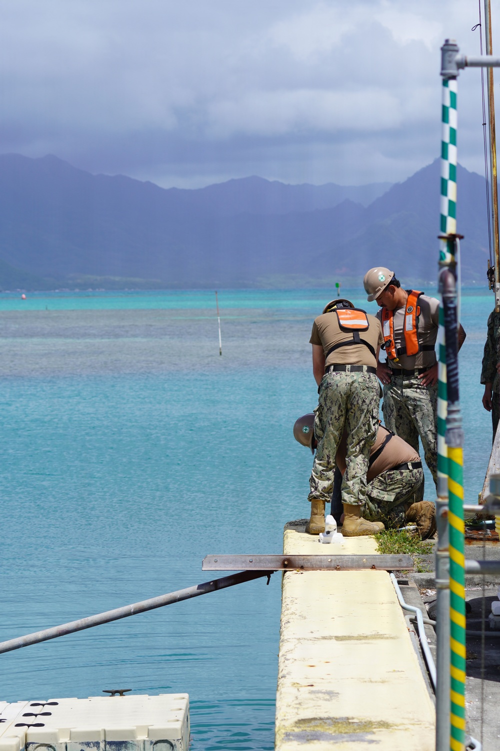 Military personnel in camouflage uniforms are working on a concrete pier or dock edge near turquoise tropical waters. At least three individuals are visible, with one wearing a hard hat and orange life vest, and another crouching near the water's edge. A green and yellow checkered navigation pole is visible on the right side of the frame, with mountainous terrain visible in the background.