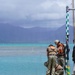 Military personnel in camouflage uniforms are working on a concrete pier or dock edge near turquoise tropical waters. At least three individuals are visible, with one wearing a hard hat and orange life vest, and another crouching near the water's edge. A green and yellow checkered navigation pole is visible on the right side of the frame, with mountainous terrain visible in the background.