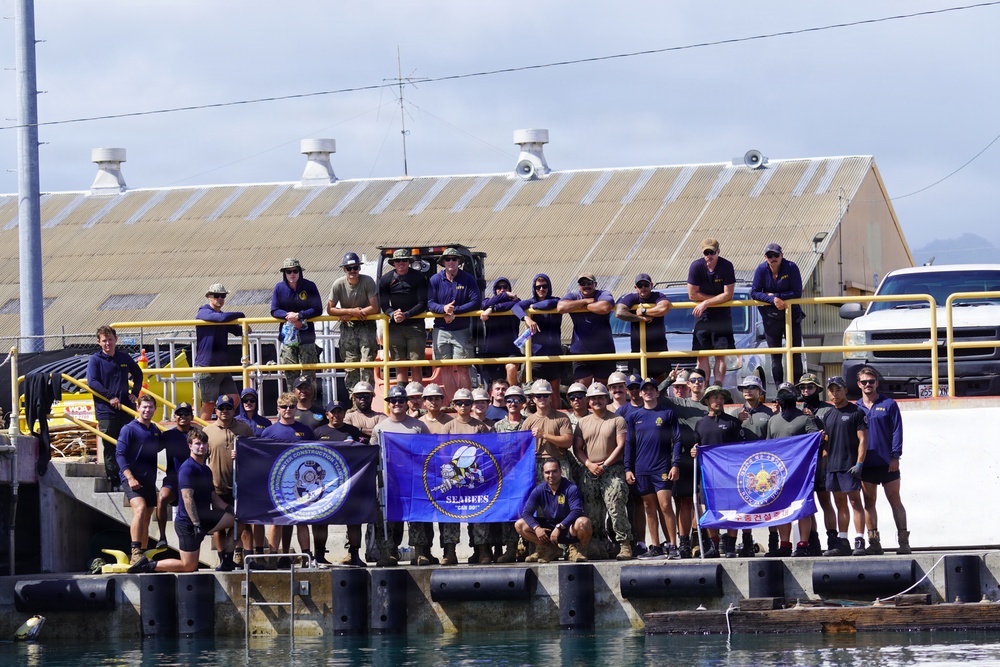 A large group of military personnel, identifiable as U.S. Navy Seabees (Naval Construction Battalion), pose for a group photo at a waterfront dock or pier facility. Personnel are arranged on two levels — a lower dock area and an upper platform with yellow safety railings. Three blue unit flags/banners are prominently displayed, with the center banner clearly reading 'SEABEES'.