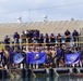 A large group of military personnel, identifiable as U.S. Navy Seabees (Naval Construction Battalion), pose for a group photo at a waterfront dock or pier facility. Personnel are arranged on two levels — a lower dock area and an upper platform with yellow safety railings. Three blue unit flags/banners are prominently displayed, with the center banner clearly reading 'SEABEES'.