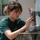 A young woman wearing a green 'CMML' t-shirt is carefully installing or adjusting a trail/wildlife camera onto a metal pole secured with a strap. She is focused intently on the device, using both hands to position or configure it. The setting appears to be an outdoor area near a building, photographed in bright daylight.