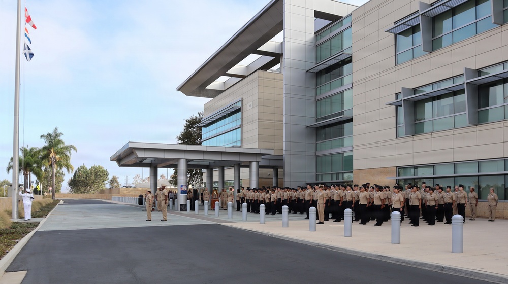 A large formation of military personnel in tan/khaki uniforms are assembled in front of a modern multi-story government or military facility. The personnel appear to be participating in a formal ceremony or muster, with officers standing at the front facing the formation. A flagpole with flags is visible on the left side of the image.