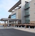 A large formation of military personnel in tan/khaki uniforms are assembled in front of a modern multi-story government or military facility. The personnel appear to be participating in a formal ceremony or muster, with officers standing at the front facing the formation. A flagpole with flags is visible on the left side of the image.