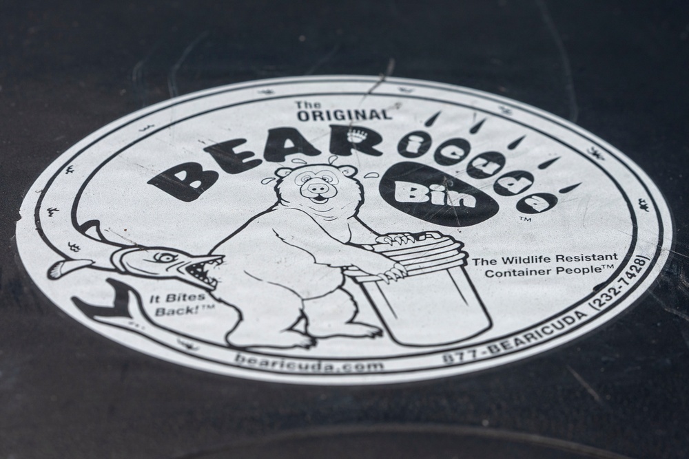 The image shows a circular black-and-white label/sticker affixed to a dark surface, belonging to a product called 'The Original BEARicuda Bin.' The label advertises a wildlife-resistant container product with cartoon illustrations of a bear and a barracuda fish. The branding includes slogans, a website, and a phone number.