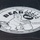 The image shows a circular black-and-white label/sticker affixed to a dark surface, belonging to a product called 'The Original BEARicuda Bin.' The label advertises a wildlife-resistant container product with cartoon illustrations of a bear and a barracuda fish. The branding includes slogans, a website, and a phone number.