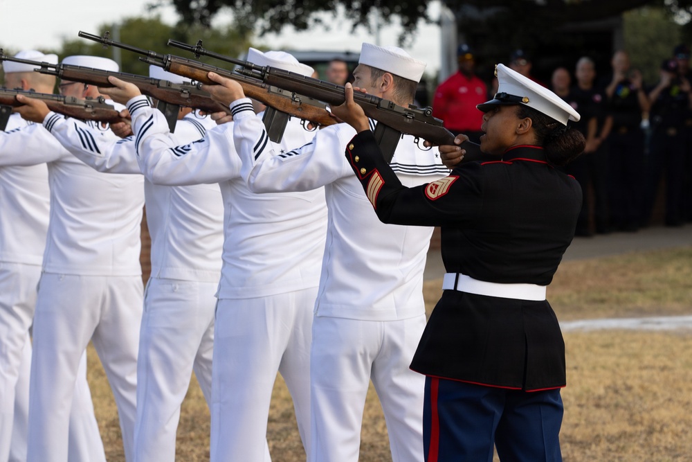 A military funeral or ceremonial honor guard is performing a rifle salute. A group of U.S. Navy sailors in white dress uniforms are firing rifles in unison, while a U.S. Marine Corps sergeant in dress blues stands at the right end of the line. Spectators and observers are visible in the background.