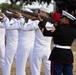 A military funeral or ceremonial honor guard is performing a rifle salute. A group of U.S. Navy sailors in white dress uniforms are firing rifles in unison, while a U.S. Marine Corps sergeant in dress blues stands at the right end of the line. Spectators and observers are visible in the background.