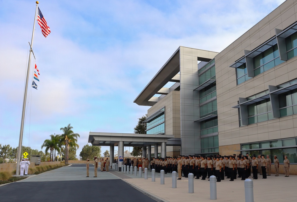 A formal military formation is taking place outside a modern multi-story building, likely a U.S. Navy installation based on the uniforms and flags visible. Personnel in tan/khaki and dark uniforms are assembled in organized ranks near the building's entrance, while two individuals in white dress uniforms stand near the flagpole. The American flag and what appears to be a Navy or command flag are displayed on a tall flagpole to the left.