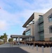 A formal military formation is taking place outside a modern multi-story building, likely a U.S. Navy installation based on the uniforms and flags visible. Personnel in tan/khaki and dark uniforms are assembled in organized ranks near the building's entrance, while two individuals in white dress uniforms stand near the flagpole. The American flag and what appears to be a Navy or command flag are displayed on a tall flagpole to the left.