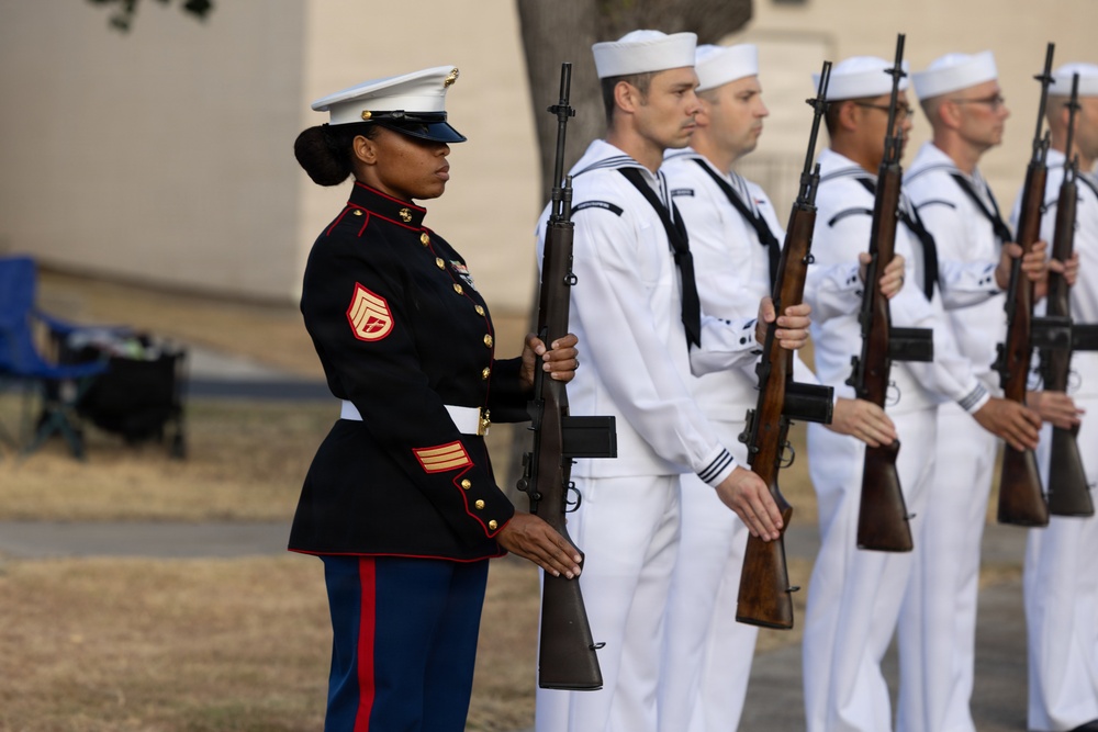 A formal military ceremony featuring a U.S. Marine Corps female staff sergeant in dress blues standing at attention alongside U.S. Navy sailors in dress whites. All personnel are holding rifles in a ceremonial position, suggesting a joint-service honor guard or parade formation. The setting appears to be an outdoor venue, likely a military installation.